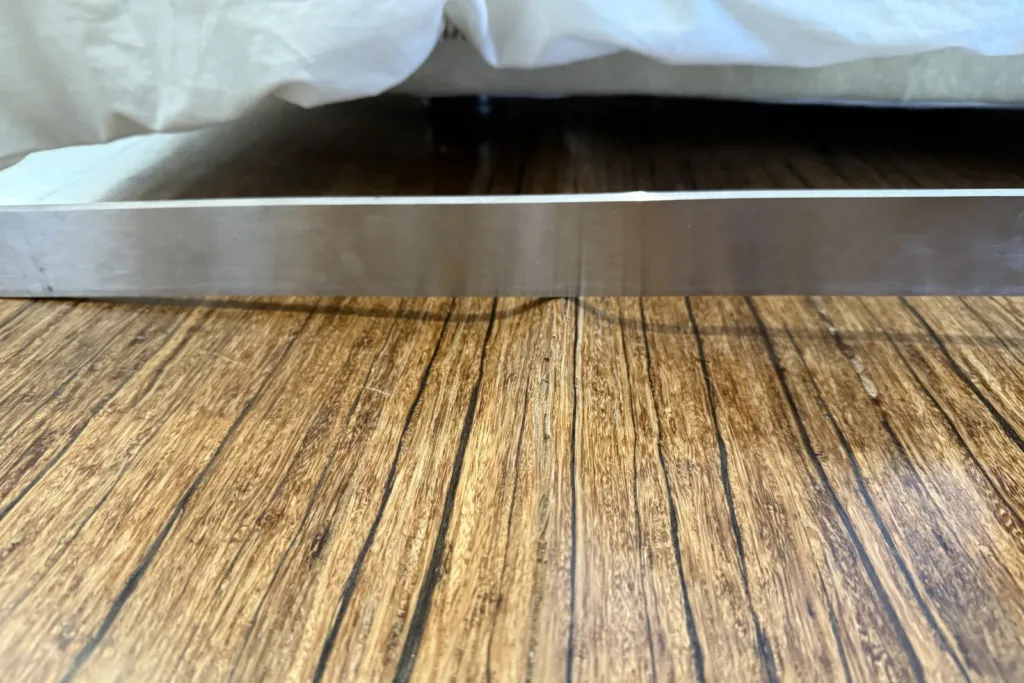 Regla metálica sobre una tarima de madera exótica abarquillada: la luz pasa bajo el centro de cada tabla y los bordes tocan la regla