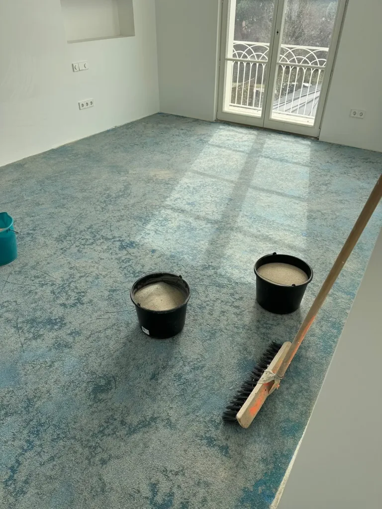 Dos cubos con arena y un cepillo sobre una solera con imprimación azul cubierta de arena