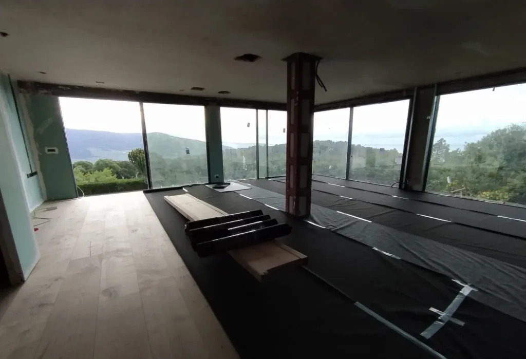 Sala acristalada con pilar metálico y vistas a la sierra: a la izquierda, tablas de roble descubiertas; el resto del suelo, cubierto con lámina negra de juntas encintadas y rollos de lámina encima