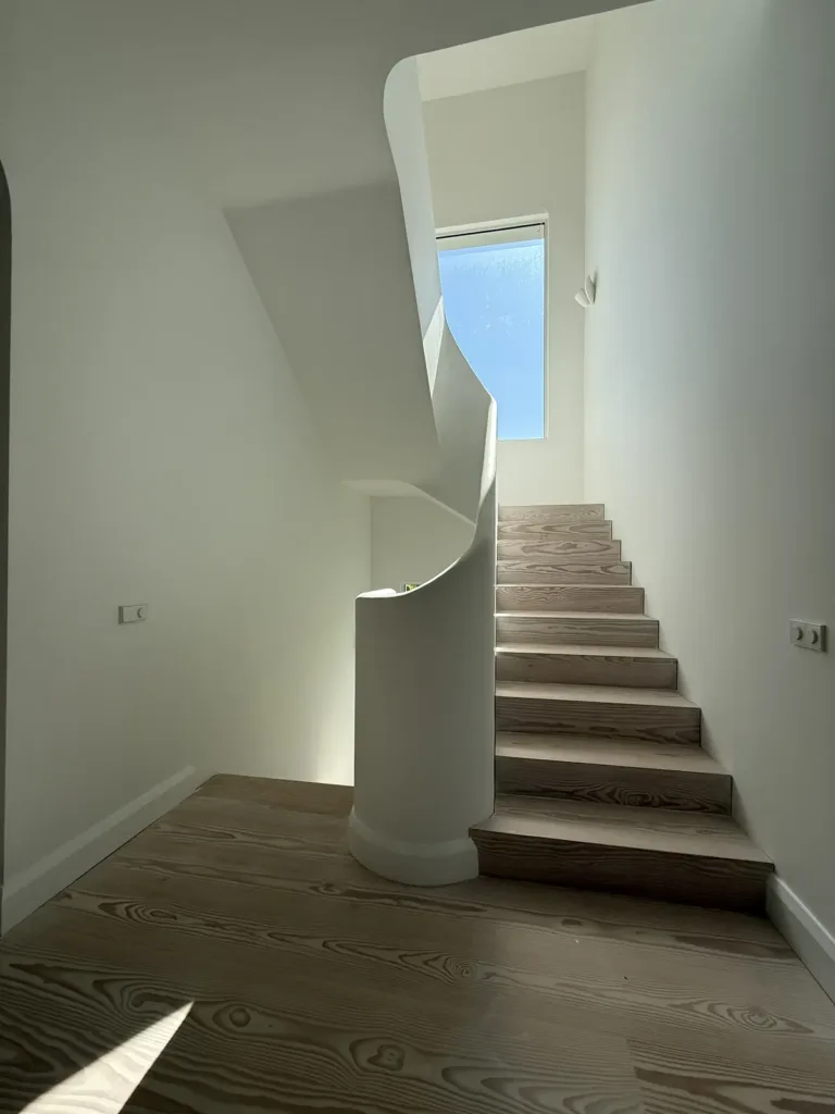 Tramo recto de escalera con huellas y tabicas de Douglas claro, entre una pared y un peto curvo de yeso que remata en un pilar redondeado
