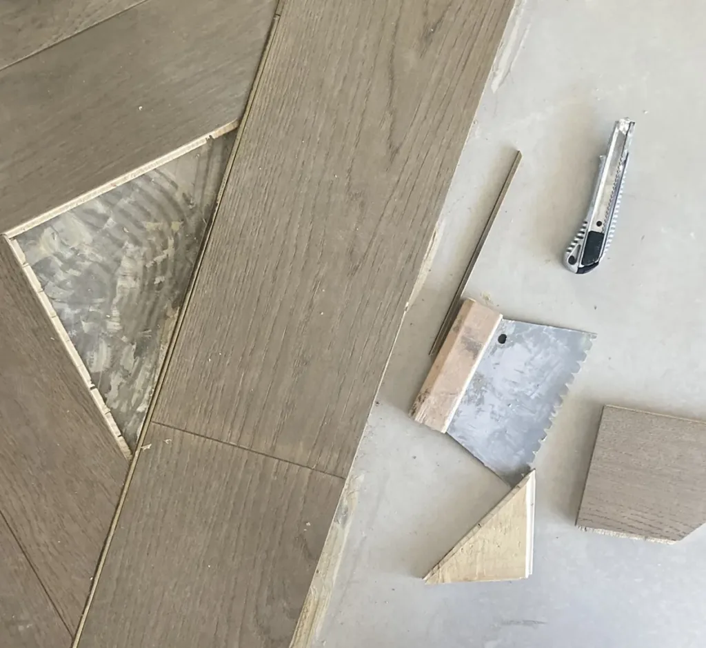 Piezas de madera de tono gris separadas por filetes metálicos dorados; un hueco triangular deja ver el adhesivo y al lado hay una pieza triangular, una llana dentada y un cúter