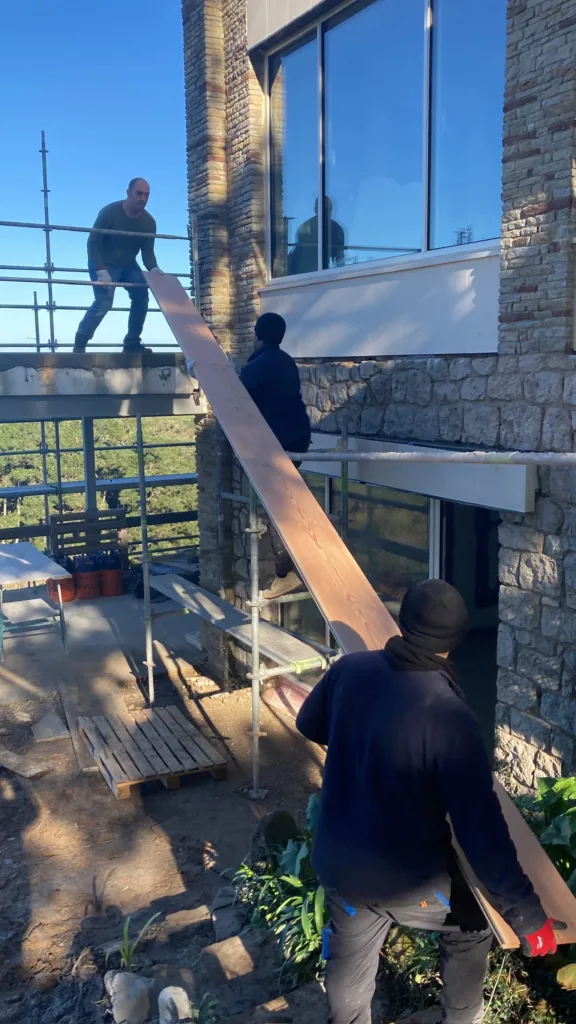 Tres operarios pasan una tabla Dinesen Douglas de gran longitud por el andamio de la fachada de piedra