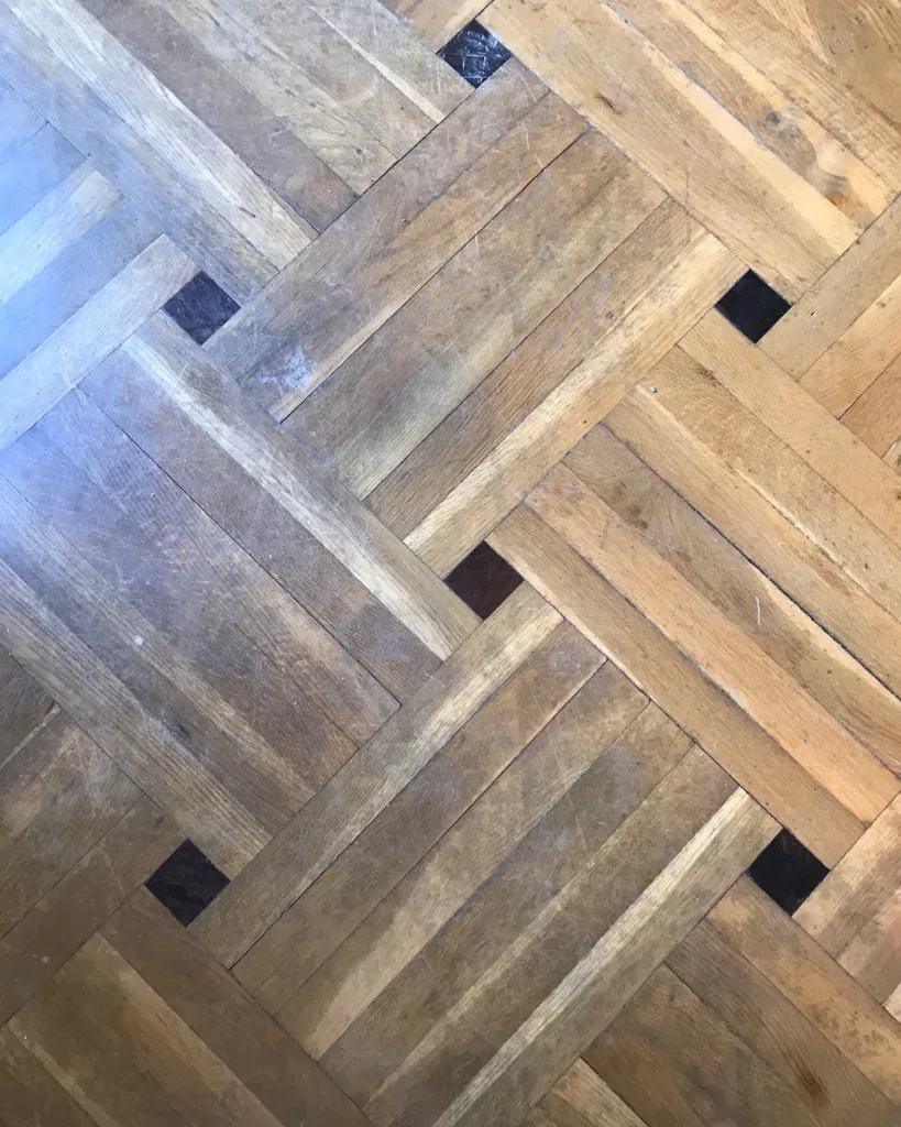 Parquet antiguo de roble en cestería con dados oscuros: acabado desaparecido en las zonas de paso, arañazos y alguna junta abierta