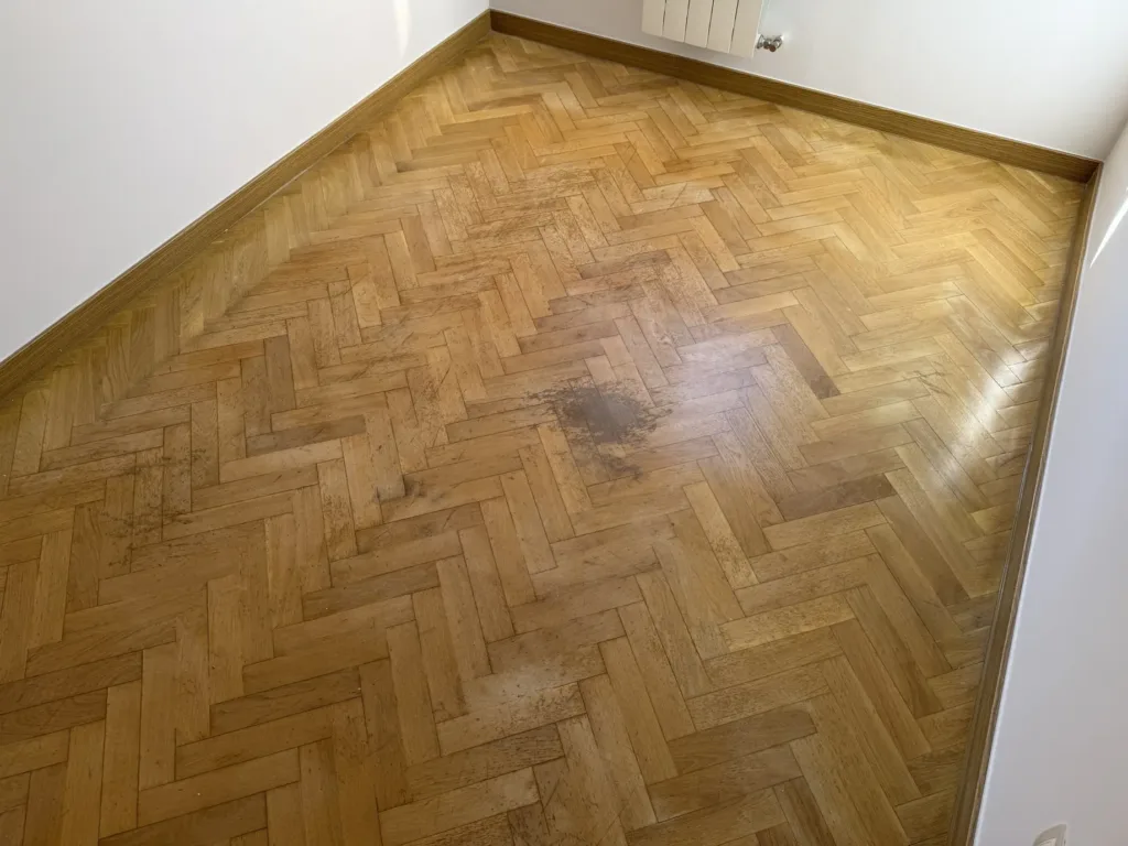 Parquet de roble en espiga antes del lijado, con arañazos y una mancha oscura en el centro de un dormitorio, piso con mirador en Madrid