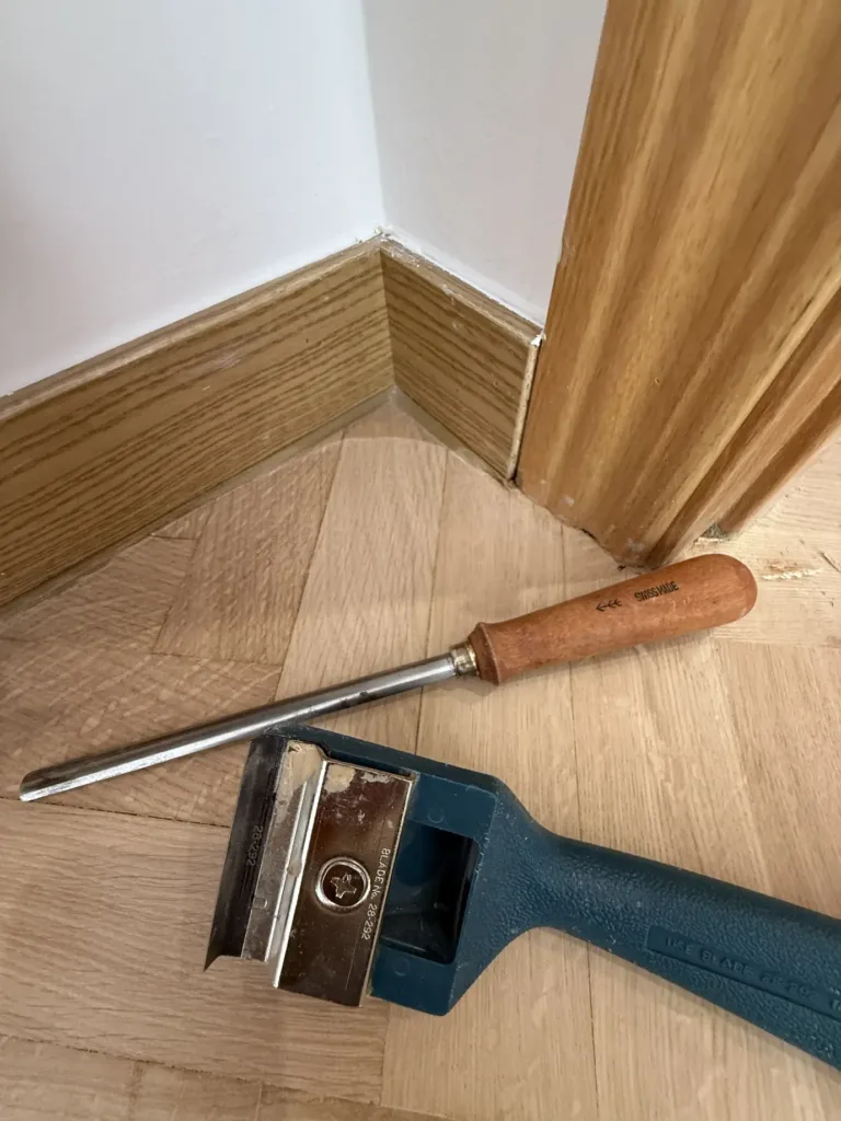 Rasqueta de cuchilla y gubia sobre el parquet de roble lijado, en el rincón entre el rodapié y el marco de la puerta, piso con mirador en Madrid