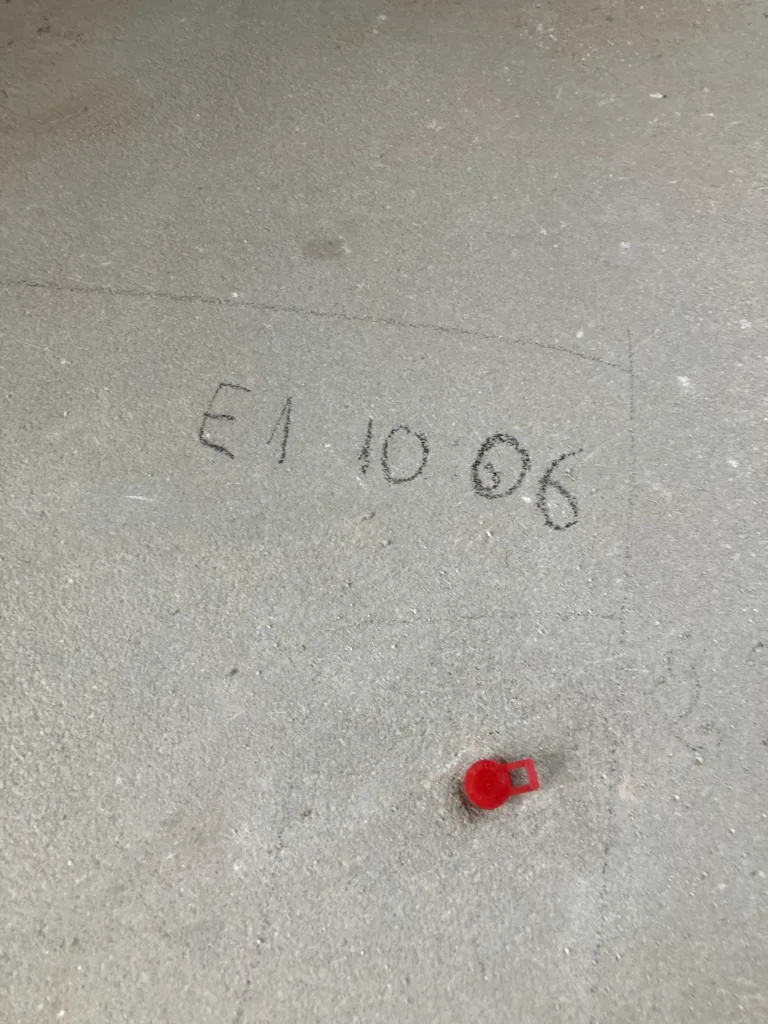 Punto de ensayo E1 marcado a lápiz en la solera, con el tapón rojo del taladro de la sonda de humedad