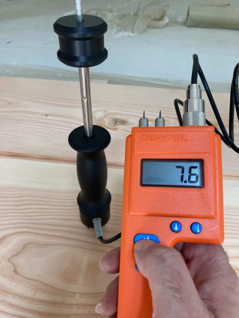 Medidor de humedad de madera con sonda de martillo marcando 7,6 % sobre una tabla Douglas