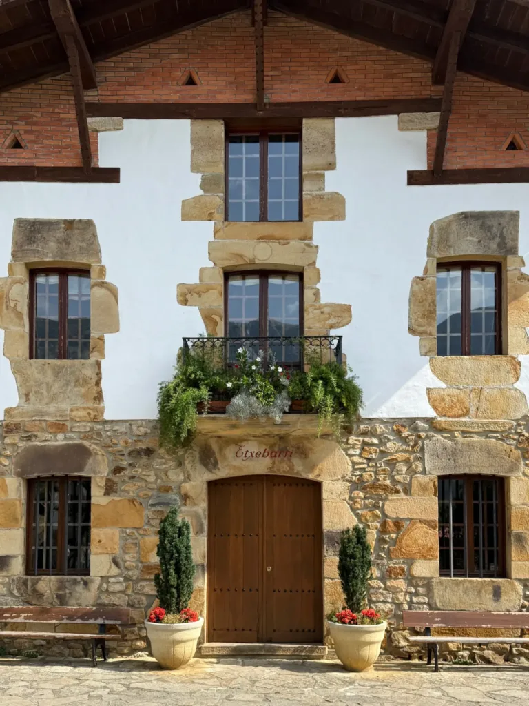 Fachada principal del Asador Etxebarri: sillería y muro encalado, balcón con plantas y puerta de madera entre dos macetas