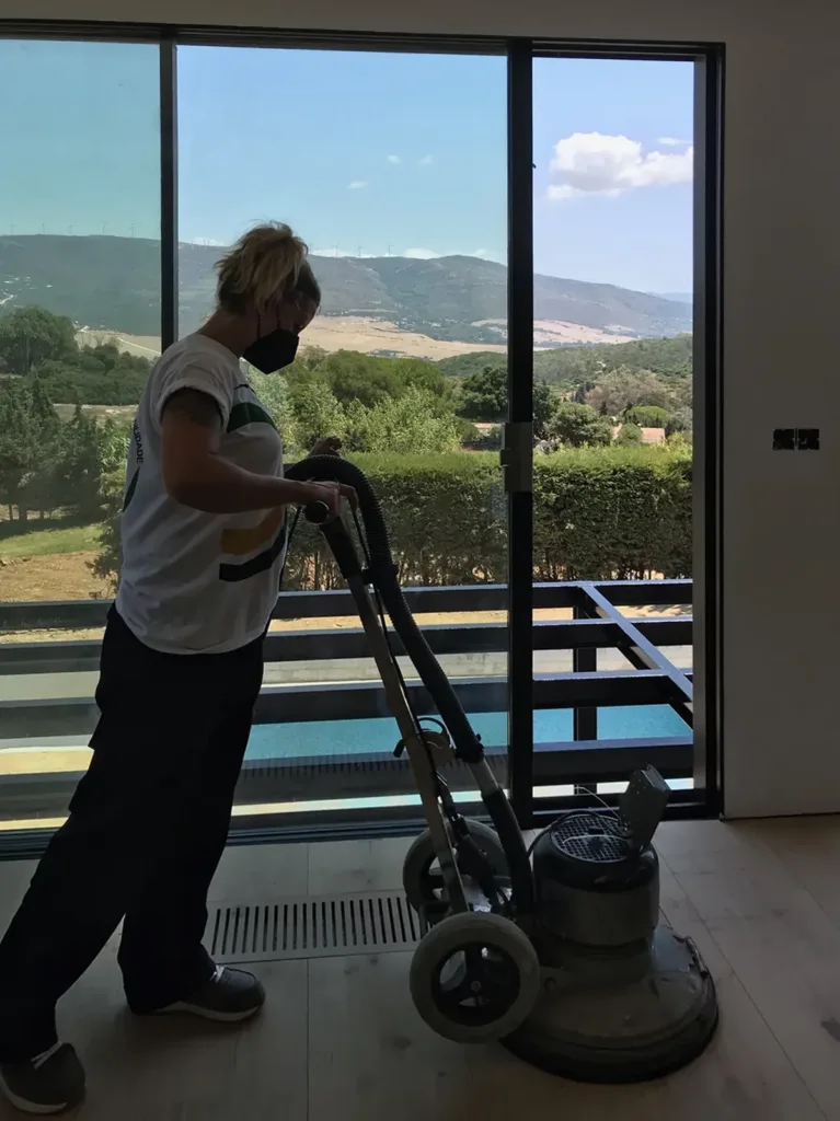 Una operaria lija con una rotativa el suelo de roble recién colocado, junto a una cristalera abierta a la sierra; a sus pies, una rejilla de clima en el suelo