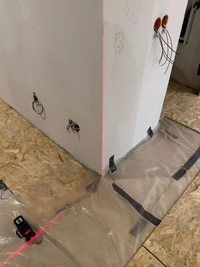 Láser de líneas sobre una lámina plástica, proyectando una línea roja por el suelo y la arista de un muro; alrededor, tableros OSB