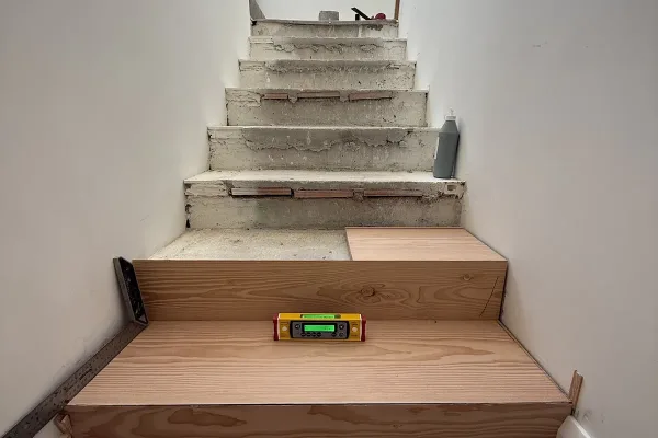 Revestir lo que ya está hecho: huella y tabica de Douglas sobre los peldaños de hormigón, comprobados con nivel Escalera de obra con los primeros peldaños ya revestidos de huella y tabica de Douglas; un nivel descansa sobre una huella y los peldaños de arriba siguen en hormigón